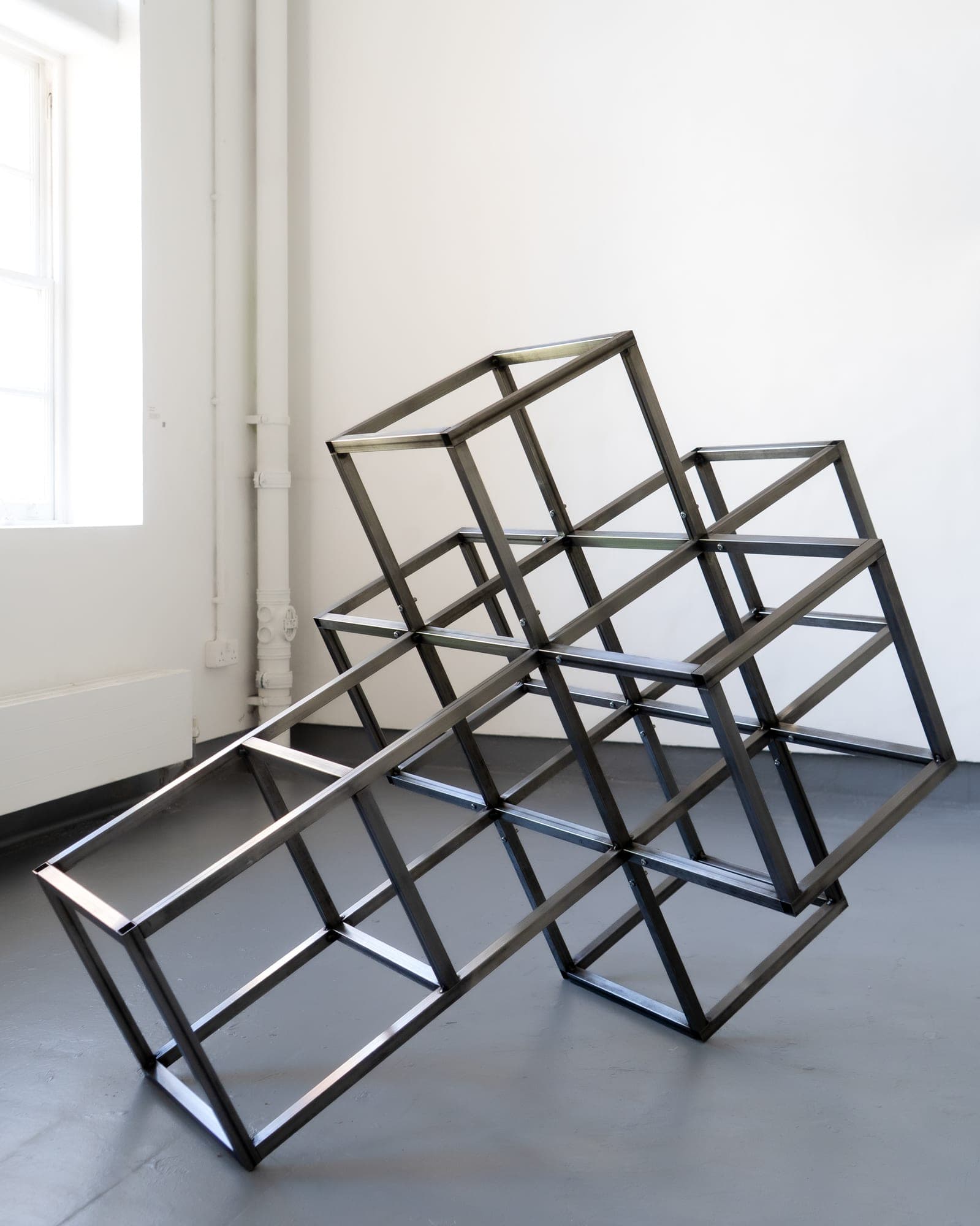 Welded steel geometric sculpture installed in a bright gallery space with a window and white wall