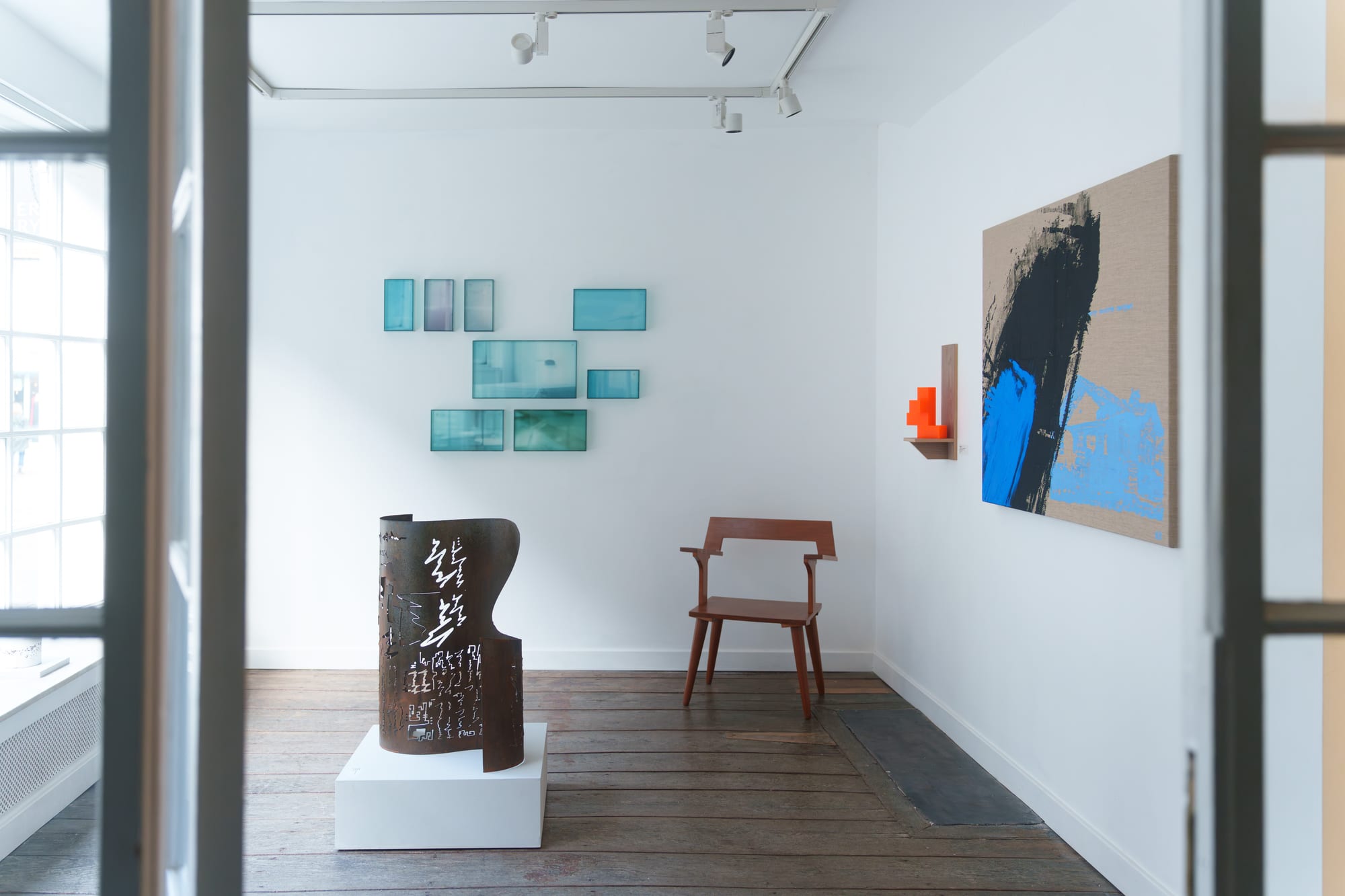 Gallery interior with wall-mounted glass panel sculptures, an orange 3D printed form, and a painting