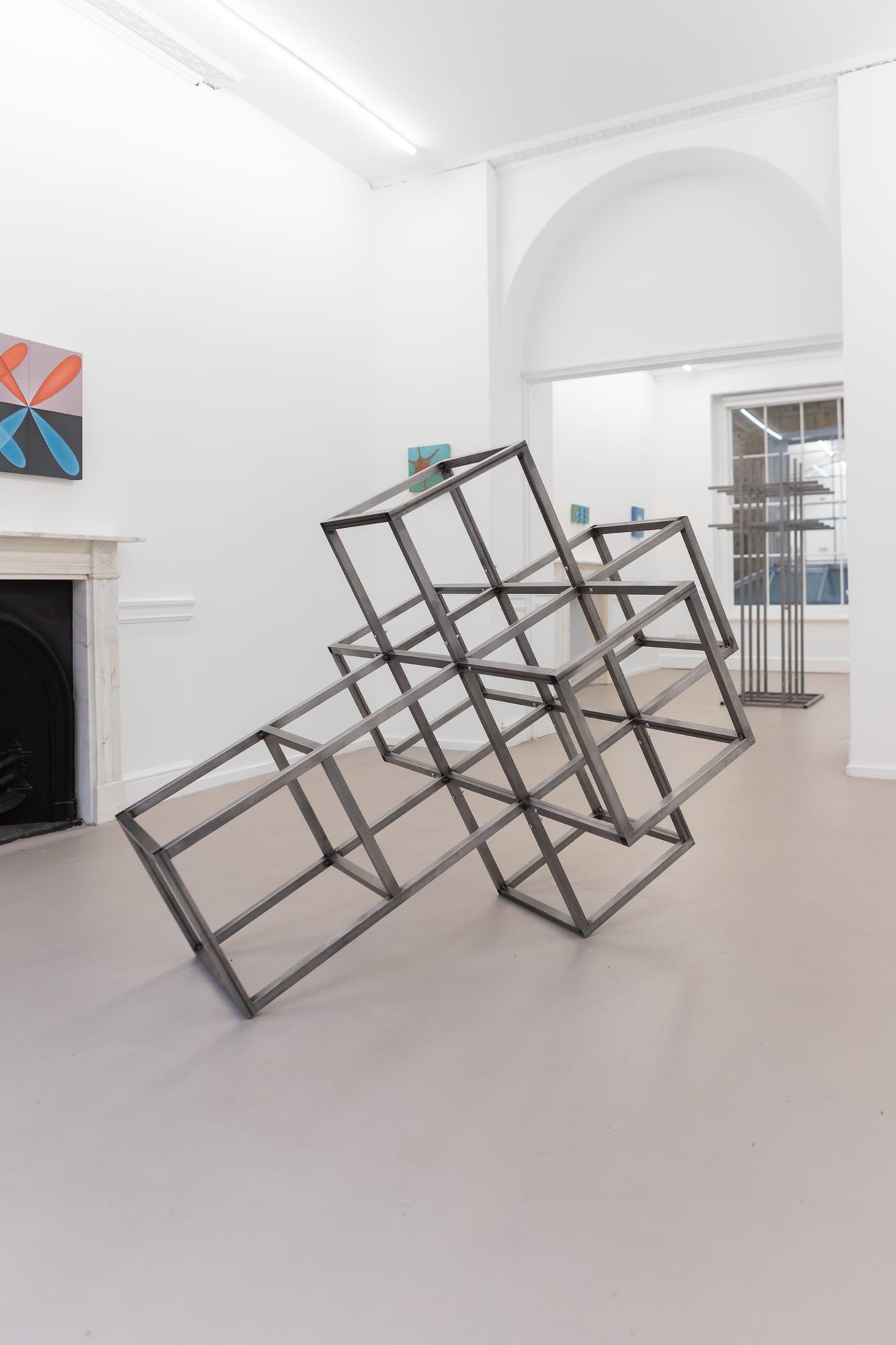 Leaning steel cube-frame sculpture in a gallery with a fireplace and paintings